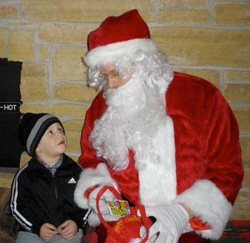 A child in a black jacket looks up at Santa Claus, who is dressed in a red suit and holding a gift.