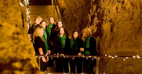 A choir of women in black outfits and green scarves performs in a dimly lit cave, decorated with string lights.