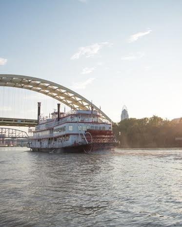 A riverboat sails under a bridge, with a city skyline visible in the background during a sunny day.