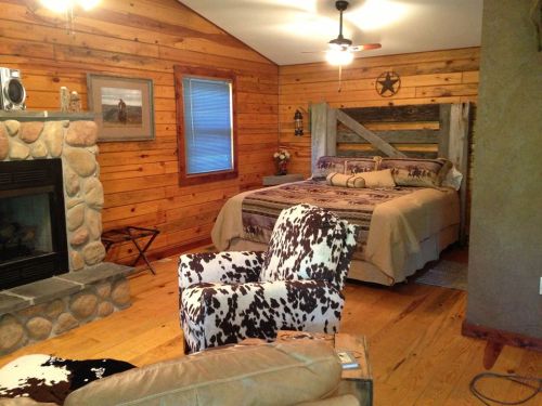 Cozy rustic bedroom with wooden walls, a stone fireplace, a large bed, and cowhide furniture.