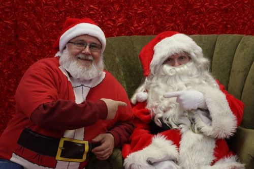 Two people dressed as Santa Claus, one in a red suit and the other in a traditional Santa outfit, sitting together and smiling.