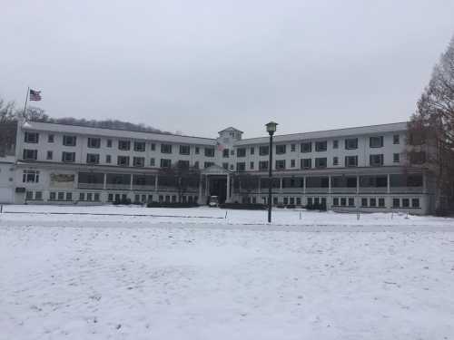 A large, white building with multiple stories, surrounded by snow, under a cloudy sky. An American flag is visible.