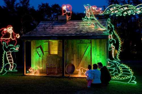 A cozy wooden cabin decorated with colorful holiday lights, featuring Santa and alligators, with two people admiring the scene.