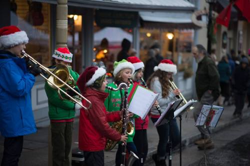 A group of young musicians in Santa hats play instruments on a festive street, with onlookers nearby.