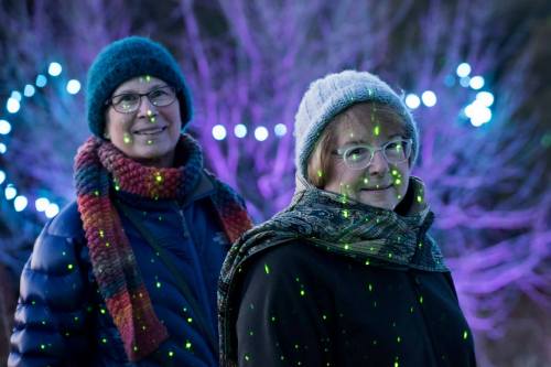 Two women stand outdoors, smiling, with colorful lights and a tree in the background, wearing winter hats and scarves.