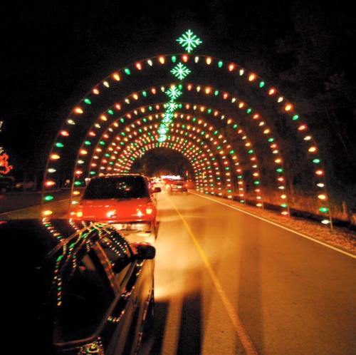 A festive light tunnel with colorful decorations, viewed from a car on a dark road.