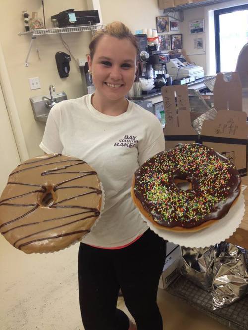 A smiling person holds two large donuts, one with chocolate icing and sprinkles, the other with a chocolate glaze.
