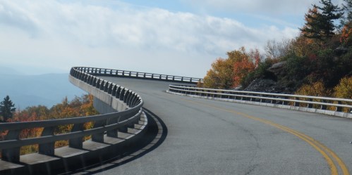 A winding road curves through a scenic landscape with colorful autumn foliage and a cloudy sky.