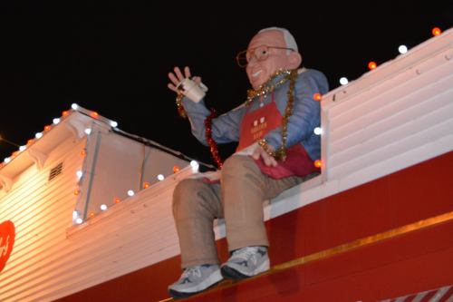 A large puppet figure wearing glasses and a blue jacket sits on a roof, adorned with festive lights and beads.