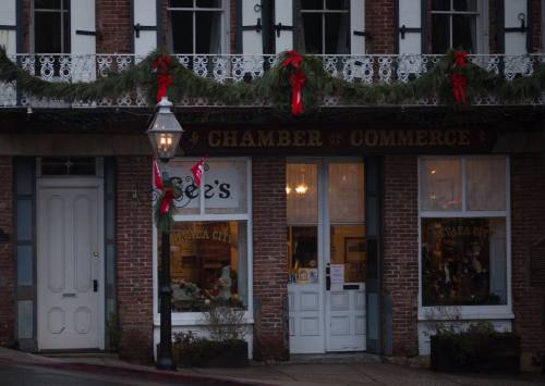 A festive storefront decorated with garlands and bows, featuring a sign for the Chamber of Commerce and a shop named "See's."