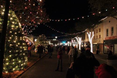 A festive street at night, adorned with colorful lights and decorated trees, with people strolling along the pathway.