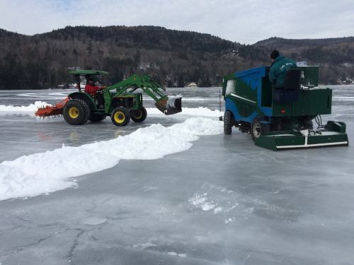 A tractor and an ice resurfacing machine clear snow on a frozen lake, surrounded by trees and a cloudy sky.