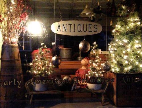 A cozy antique shop window decorated with small Christmas trees, lights, and vintage ornaments.