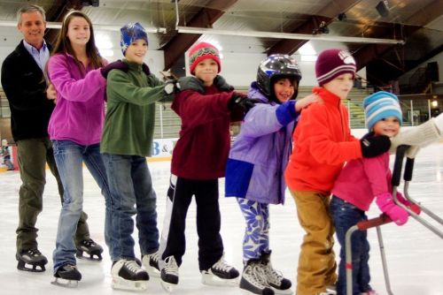 A group of children and an adult ice skating in a line, smiling and wearing colorful winter clothing and skates.
