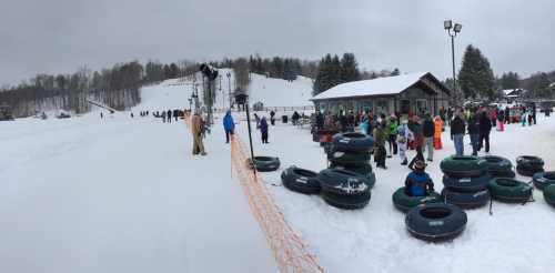 A snowy landscape with people waiting in line for tubing, surrounded by trees and a ski lift in the background.