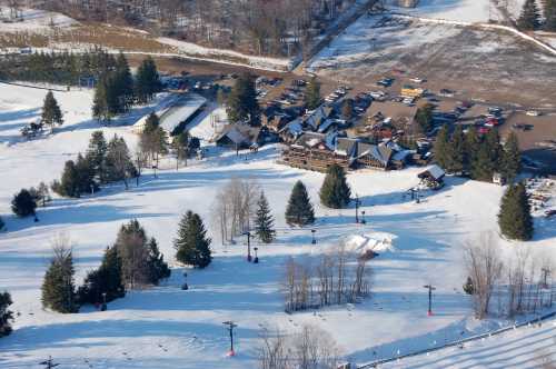 Aerial view of a snowy landscape with a ski resort, parking lot, and trees in a winter setting.