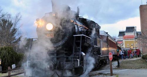 A vintage steam train billows smoke as it approaches a stop sign, with a crowd waiting nearby.