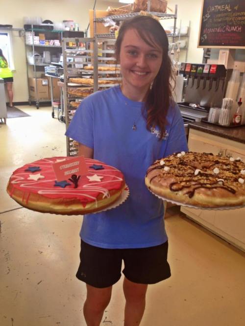 A young woman in a blue shirt holds two large decorated cakes in a bakery setting.