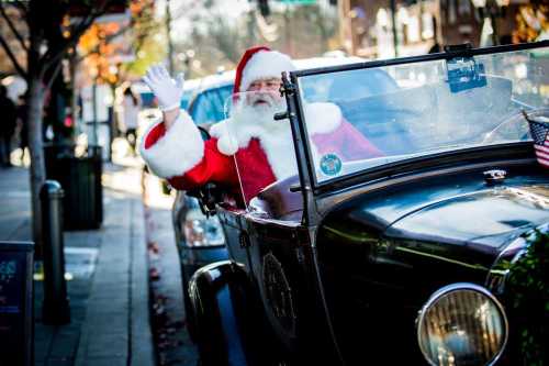 Santa Claus waves from an old-fashioned car, surrounded by a festive street scene.