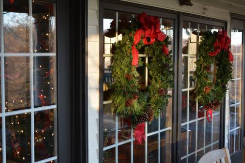 Two festive wreaths adorned with red bows and pinecones hang on windows, with holiday lights visible inside.