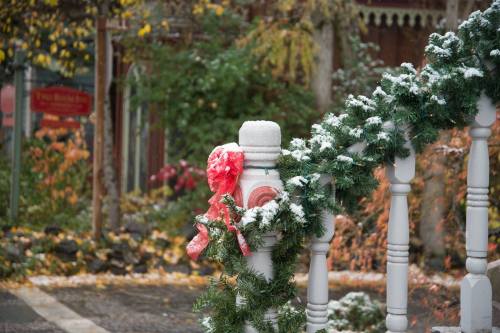 A snowy railing adorned with green garland and a red bow, surrounded by autumn foliage in a quaint outdoor setting.