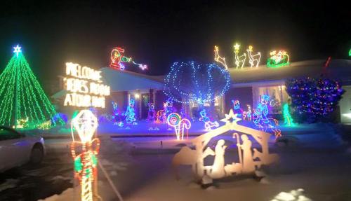 A festive house decorated with colorful Christmas lights, featuring a tree, reindeer, and a nativity scene in the snow.