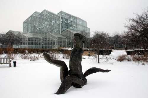 A snow-covered sculpture with abstract forms in a garden, set against a glass greenhouse in winter.