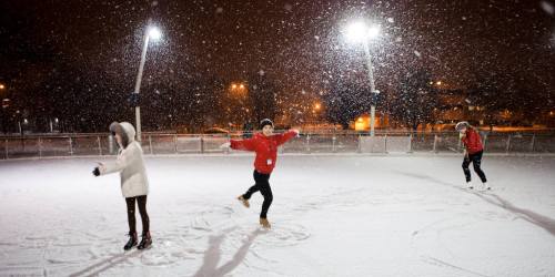 Three people ice skating in a snowy outdoor rink at night, illuminated by bright lights.