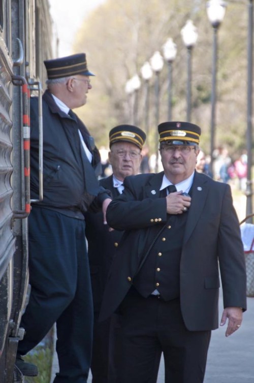 Three train conductors in uniforms stand near a train, with one adjusting his tie and others observing.