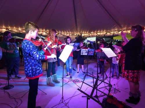 A group of young musicians playing string instruments under a tent, with colorful lights and music stands.