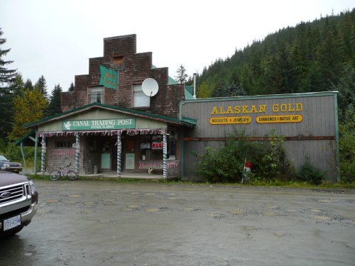 A rustic trading post with a wooden facade, surrounded by trees, featuring signs for "Canal Trading Post" and "Alaskan Gold."