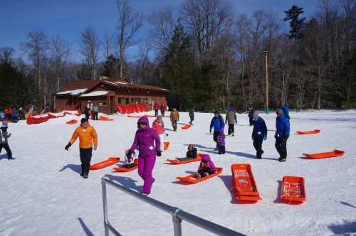 People in winter clothing walking on snow, with orange sleds scattered around and a cabin in the background.