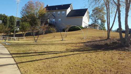 A two-story house on a grassy hill, surrounded by trees and a clear blue sky. Shadows stretch across the lawn.