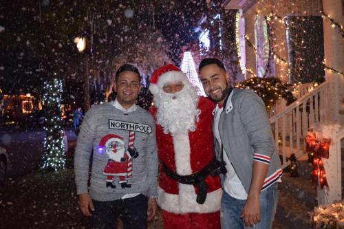 Two men pose with Santa Claus in a snowy, festive setting, surrounded by holiday lights and decorations.