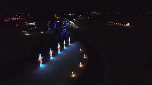Aerial view of a festive display with colorful lights, including trees and illuminated figures, set in a dark landscape.
