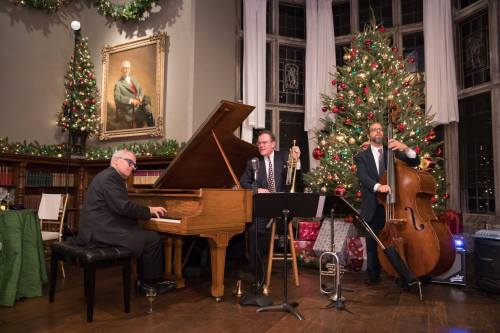 A jazz trio performs in a festive room decorated for Christmas, with a grand piano, trumpet, and double bass.