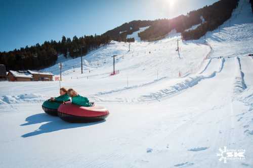 Two children ride down a snowy hill on inflatable tubes, surrounded by trees and ski lifts under a bright sun.