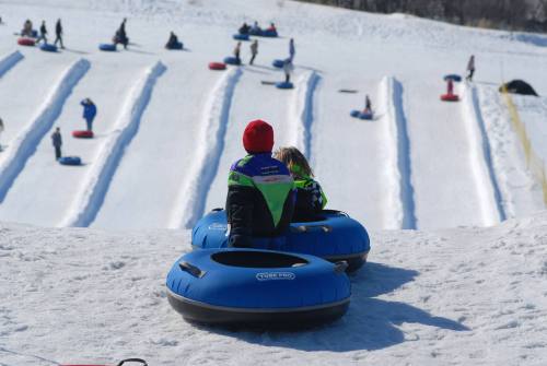 Two children sit in blue snow tubes at the top of a snowy hill, ready to slide down while others enjoy the slopes.