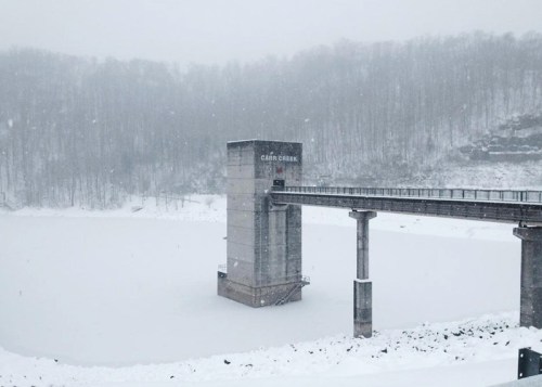 A snowy landscape featuring a dam tower over a frozen lake, surrounded by trees in a winter setting.