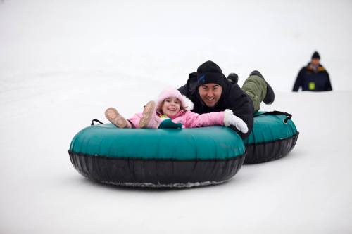 A man and a girl in winter clothing joyfully slide down a snowy hill on inflatable tubes.