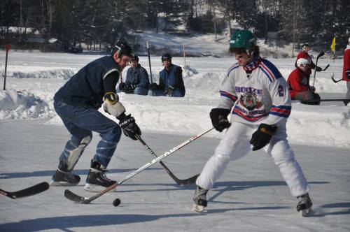 Two hockey players compete on an outdoor rink, surrounded by spectators in a snowy landscape.