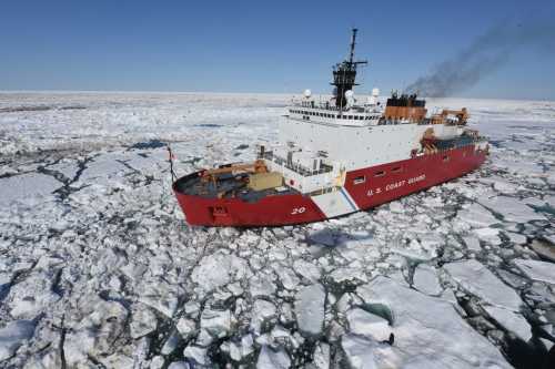 A U.S. Coast Guard icebreaker ship navigates through a sea of ice and snow under a clear blue sky.