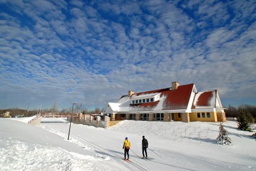 Two people ski on a snowy path near a large building under a blue sky with fluffy clouds.
