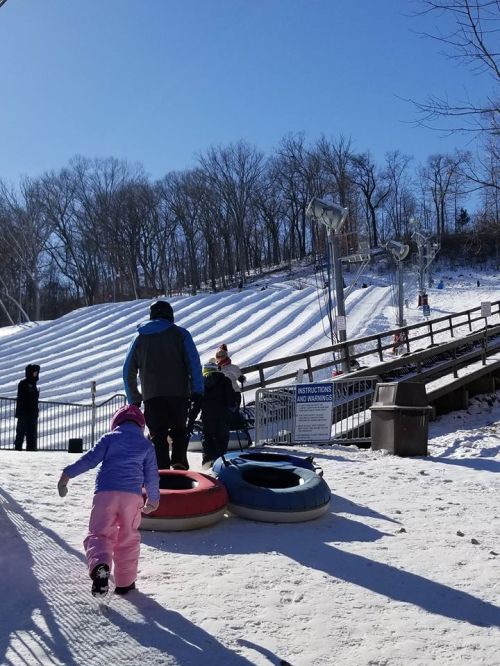 A child in winter clothing walks towards a snow tubing hill, with people tubing in the background and trees in the distance.