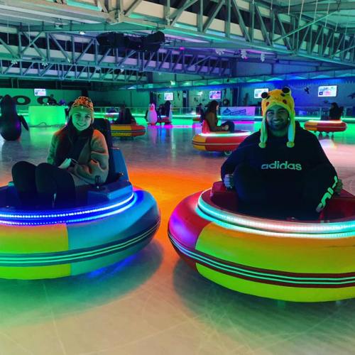 Two people in colorful bumper cars on an illuminated ice rink, wearing playful hats and smiling.