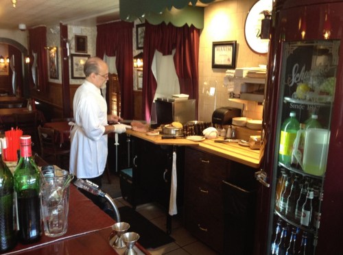 A chef prepares food in a cozy restaurant kitchen, with bottles and kitchen equipment visible in the background.