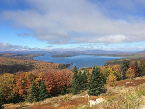 A scenic view of a lake surrounded by vibrant autumn foliage and distant mountains under a partly cloudy sky.