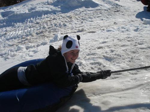 A girl in a panda hat smiles while being pulled on a snow tube across a snowy landscape.