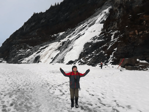 A child in a red and gray jacket stands in snow, with a waterfall and mountains in the background.