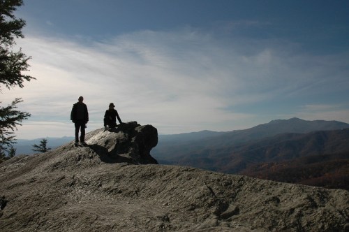 Two hikers stand on a rocky outcrop, overlooking a vast mountain landscape under a clear blue sky.